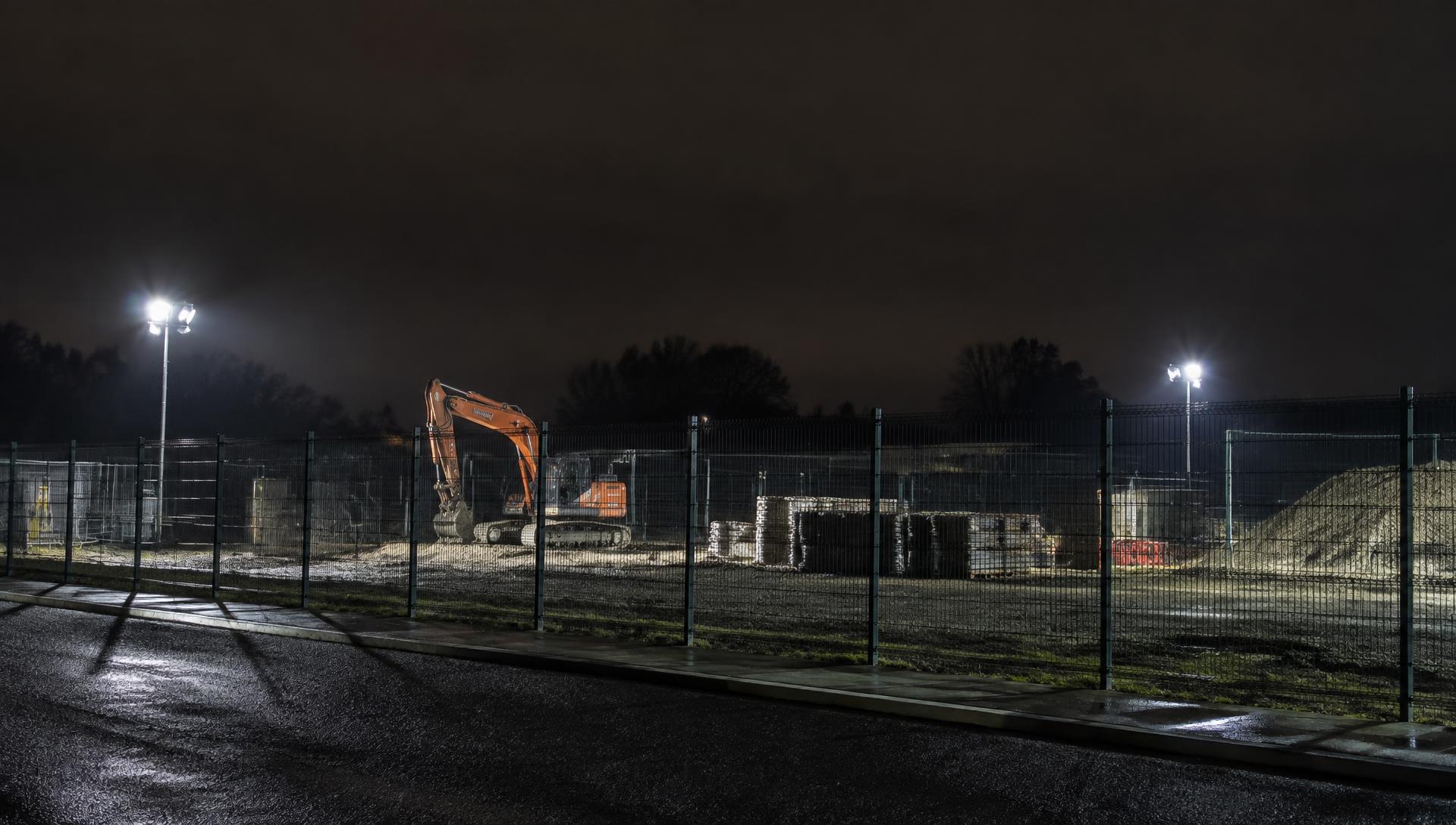 A floodlit construction site at night with plant and materials behind perimeter fencing
